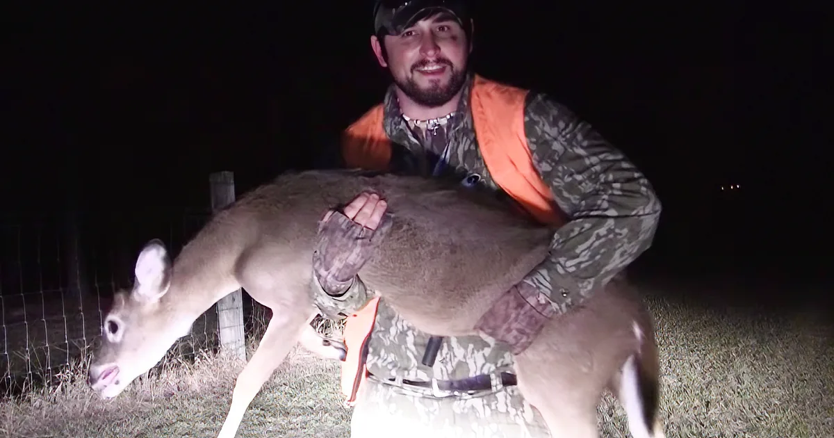 A man in camouflage holds a deer, both illuminated by a light source at night.