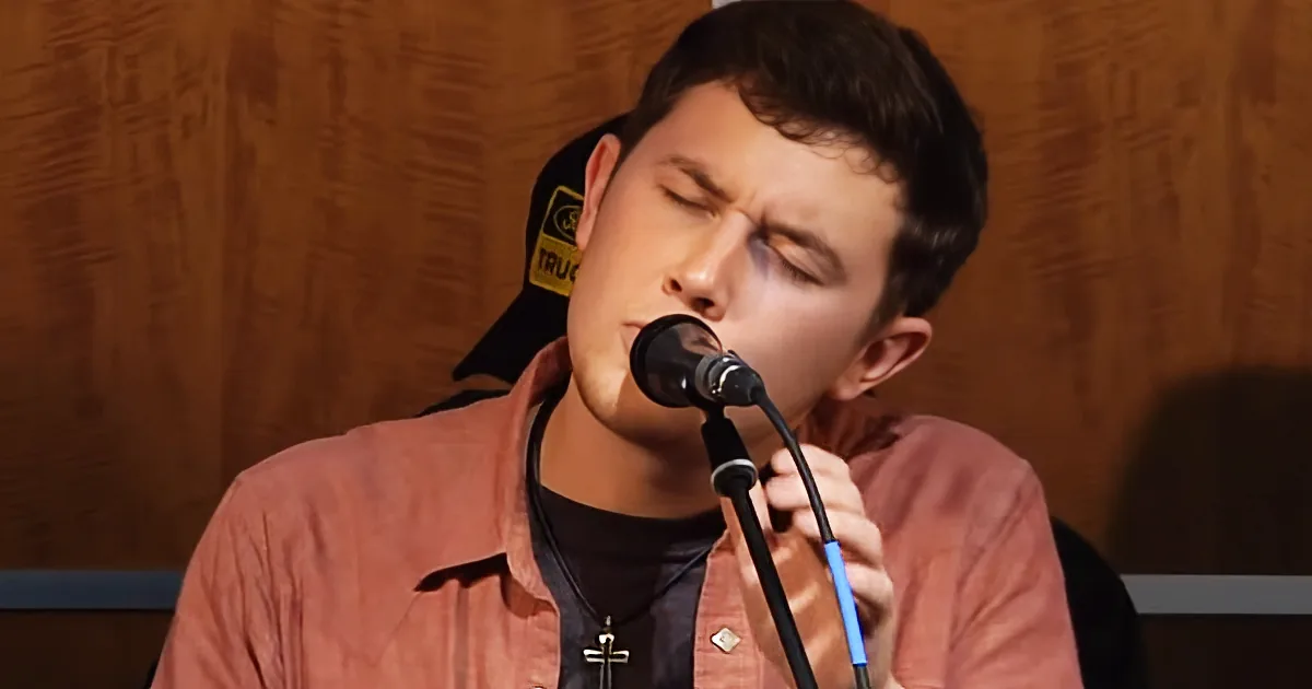 A person in a red shirt sings into a microphone, wearing a cross necklace, with a wooden backdrop.