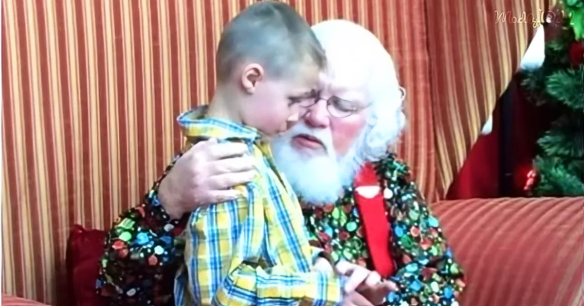 A person dressed as Santa Claus is hugging a child in a yellow plaid shirt, both seated on a red couch.