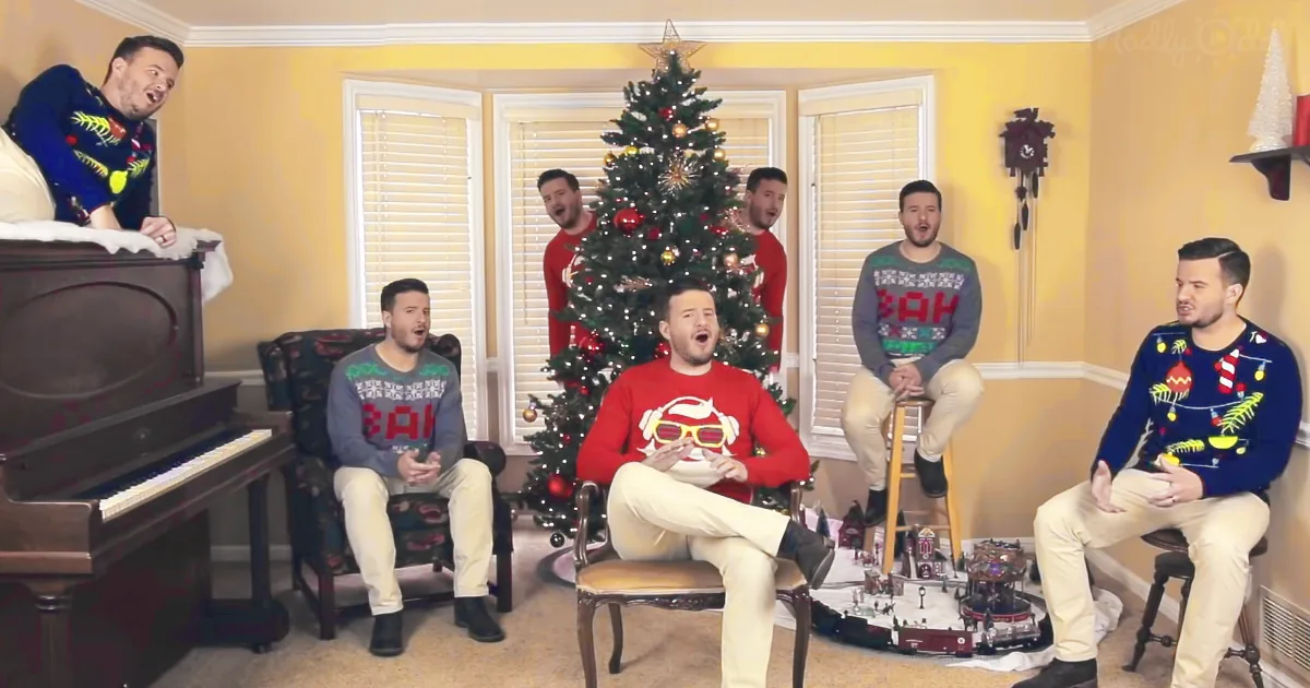 Six men in festive sweaters sing around a Christmas tree in a cozy room.