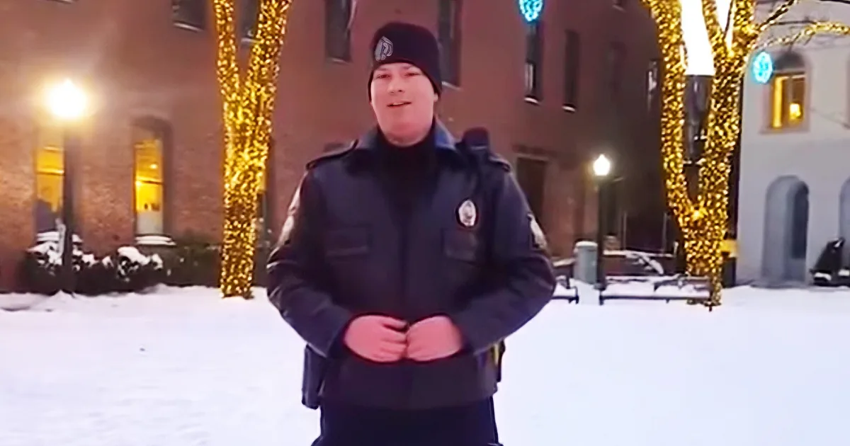 A person in a dark uniform stands in a snowy area with illuminated trees and buildings in the background.