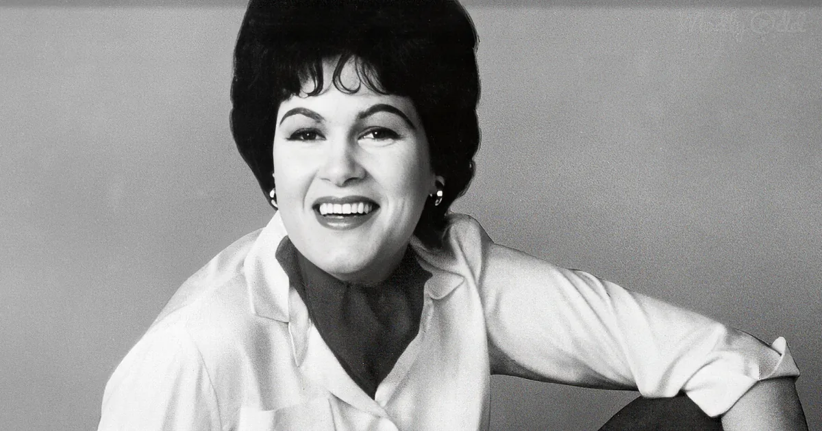 A smiling woman with short hair, wearing a white blouse, leans on a chair against a plain background.