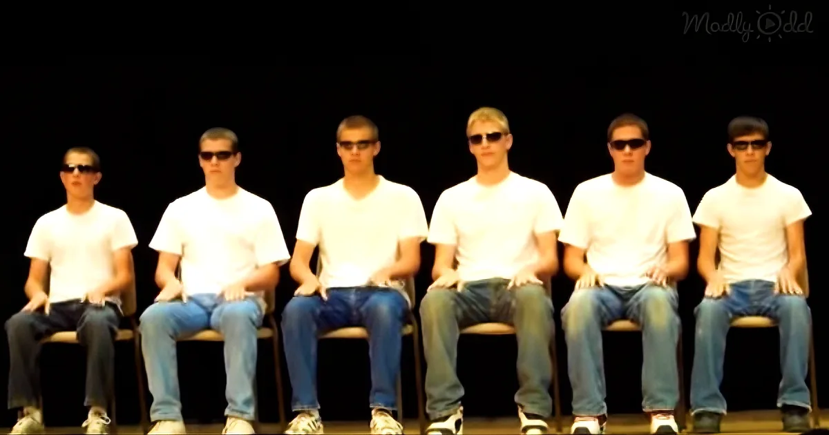 Six identical men in white shirts and jeans sit on chairs against a black background.