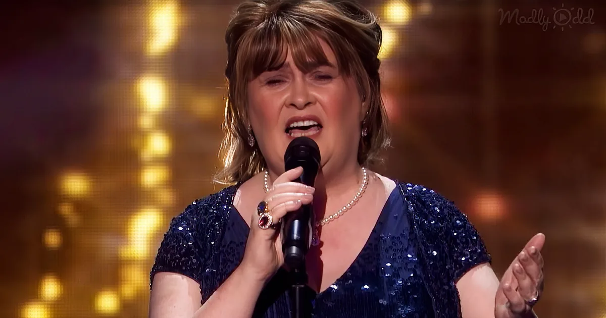 A woman in a blue sequined dress sings into a microphone, adorned with a pearl necklace and ring, against a golden backdrop.