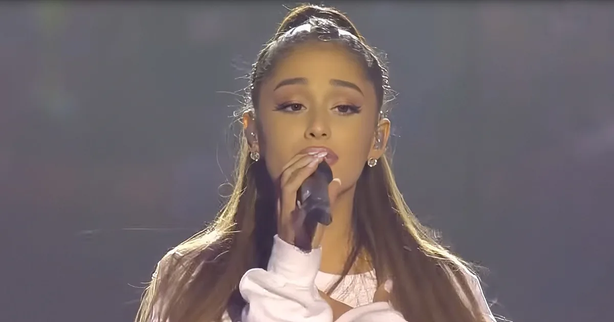 A woman with long hair, wearing a white top, sings into a microphone on stage.