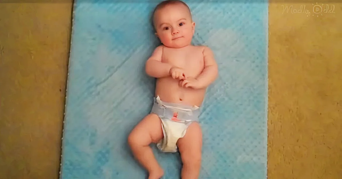 A baby in a diaper lies on a blue mat, arms crossed, looking up.
