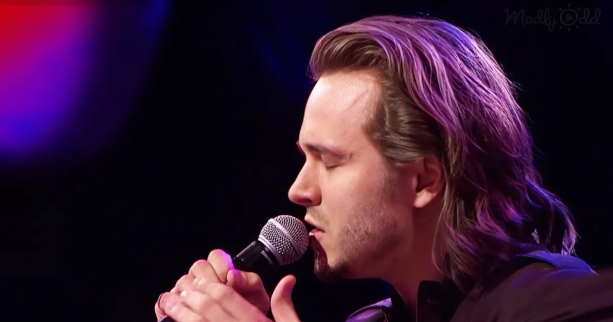 A man with long hair and a beard sings into a microphone, illuminated by purple stage lights.