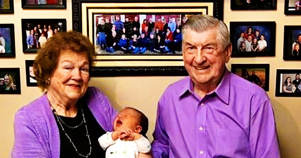 An elderly couple holds a newborn baby, surrounded by family photos on a wall.