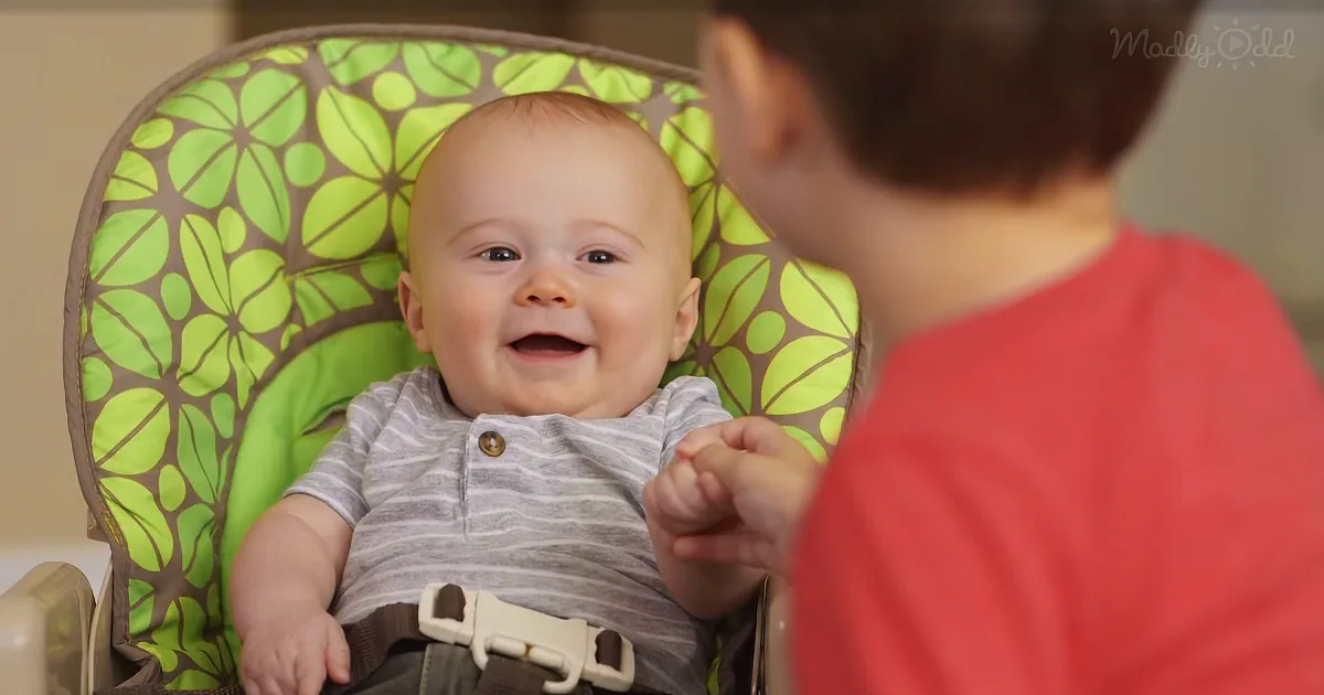 A smiling baby in a green floral high chair holds hands with a child in a red shirt.