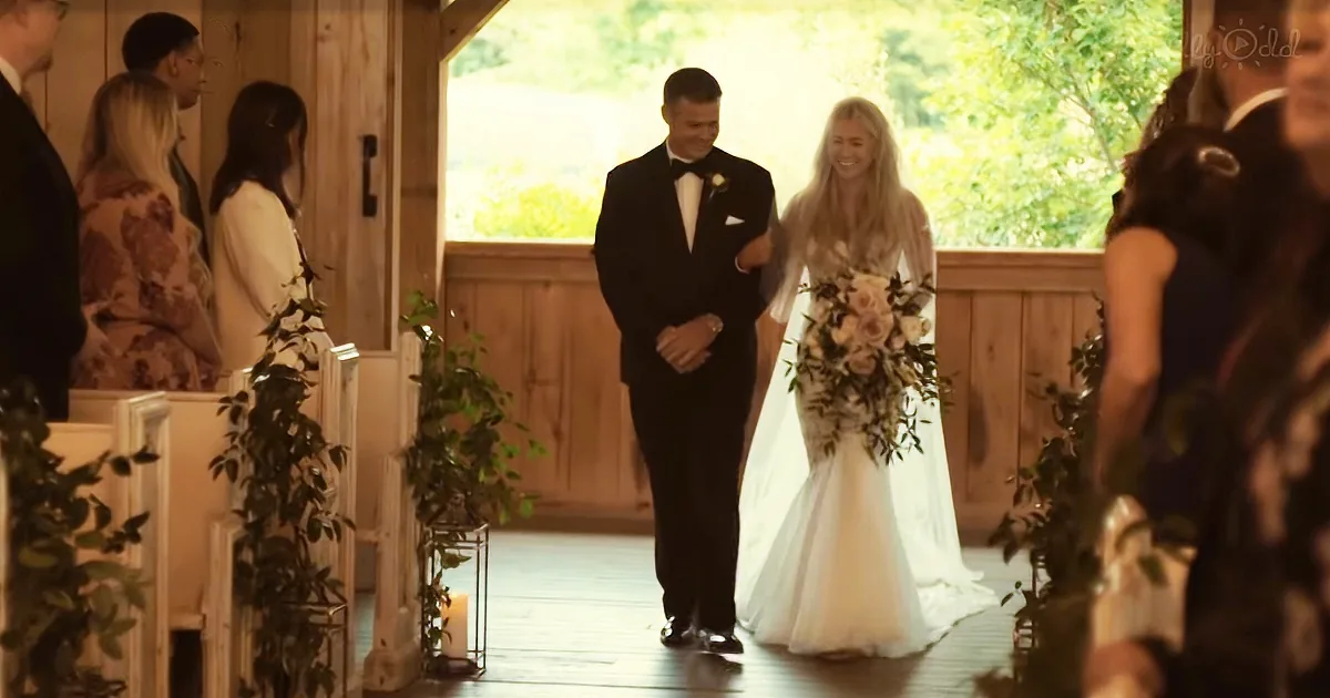 A bride in a white dress walks down the aisle with her father, holding a bouquet.