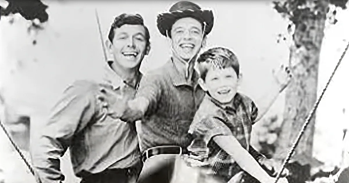 A man in a cowboy hat and two boys, one holding a stick, smile and pose together outdoors.
