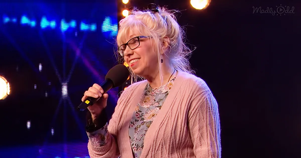 A woman with blonde hair and glasses smiles while holding a microphone on stage.