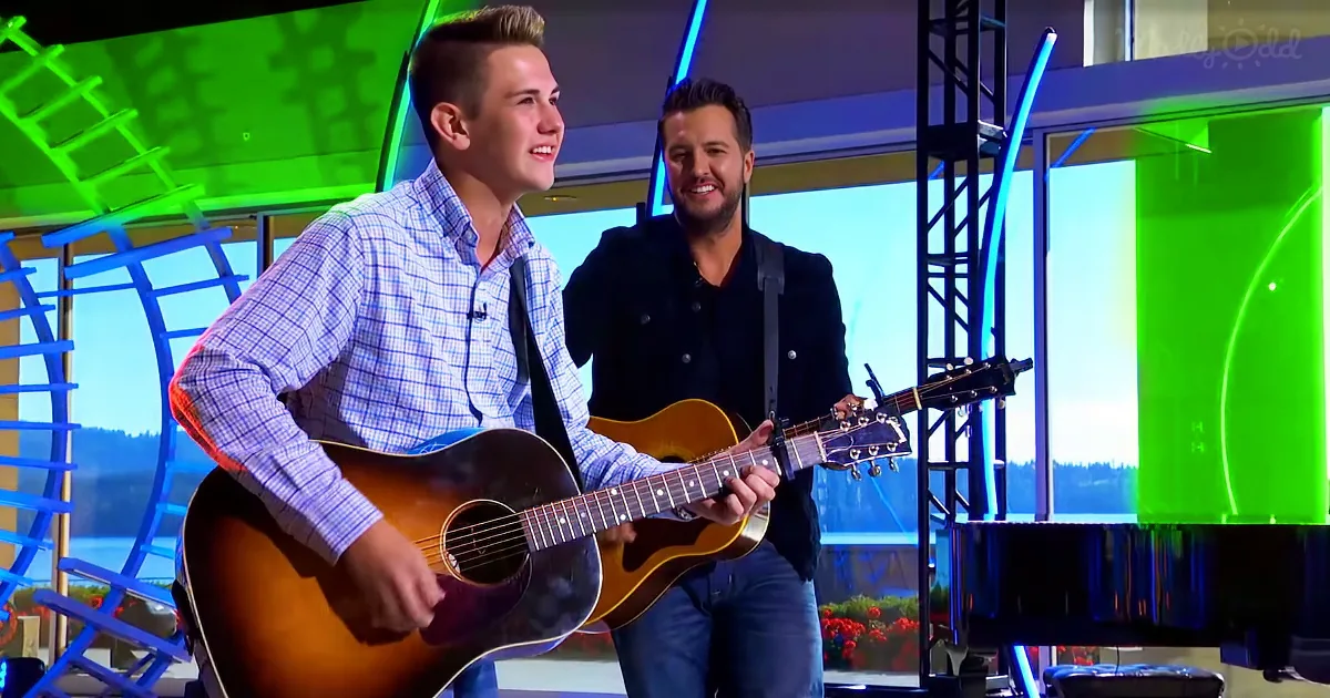 A man in a plaid shirt plays an acoustic guitar on stage, smiling, with another man in a black jacket standing behind him.