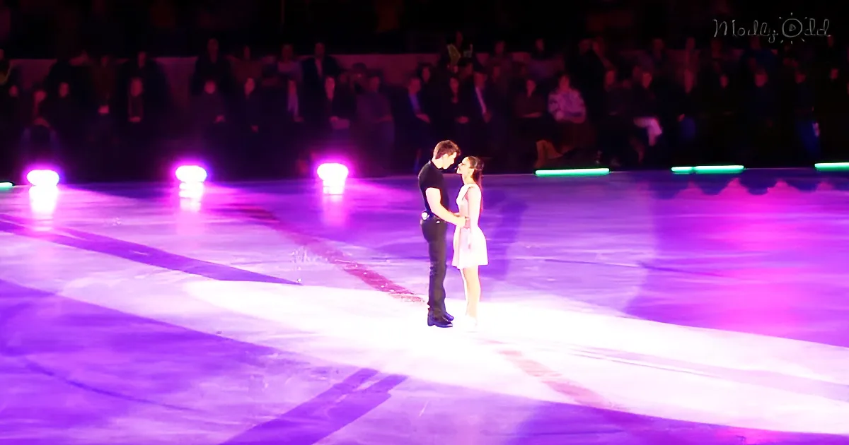 A couple stands closely on an ice rink, illuminated by purple lights, with spectators in the background.