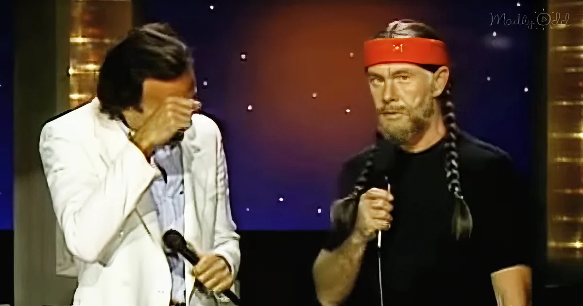 A man in a white suit holds a microphone, while another man in a black outfit with a red headband and braids speaks into...