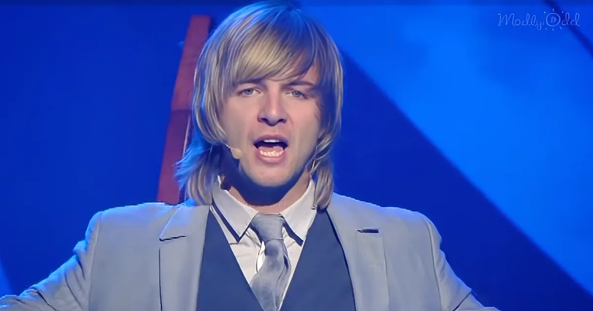 A man in a light suit and tie is singing on stage with a microphone, against a blue background.