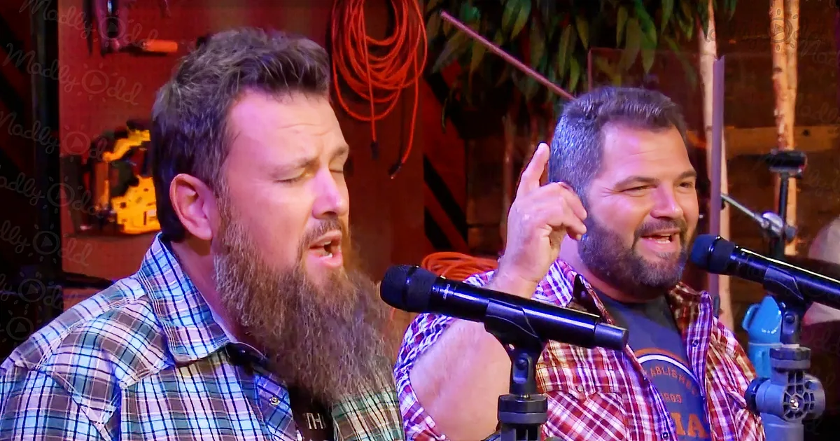 Two men with beards sing into microphones, one pointing, in a rustic setting with tools and red cords.