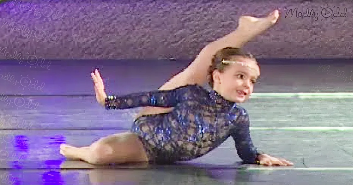 A young girl in a blue leotard performs a split on a stage with a purple background.