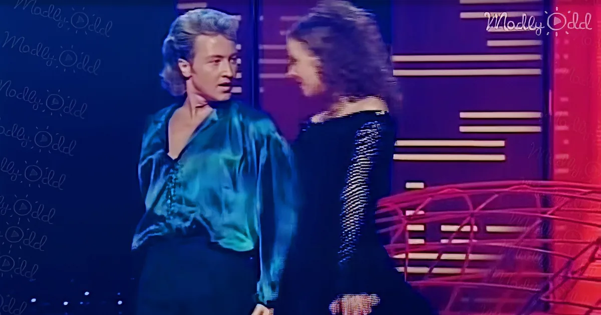 A man in a teal shirt and a woman in a black dress are facing each other on a stage with a red and blue background.