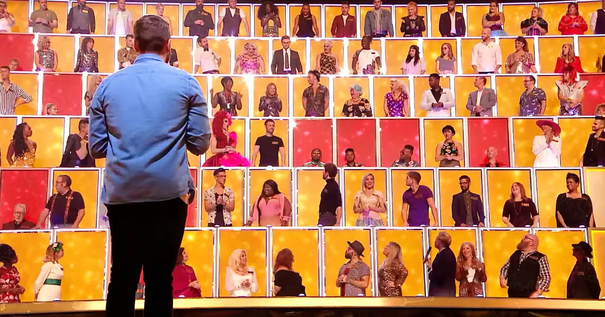 A man in a blue shirt stands on stage facing a large screen displaying multiple people in various outfits.