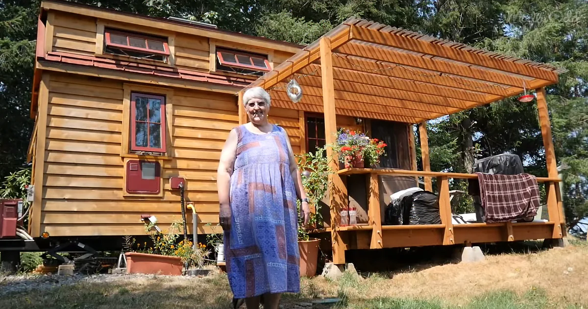 An elderly woman in a blue dress stands in front of a wooden tiny house with a covered porch and potted flowers.