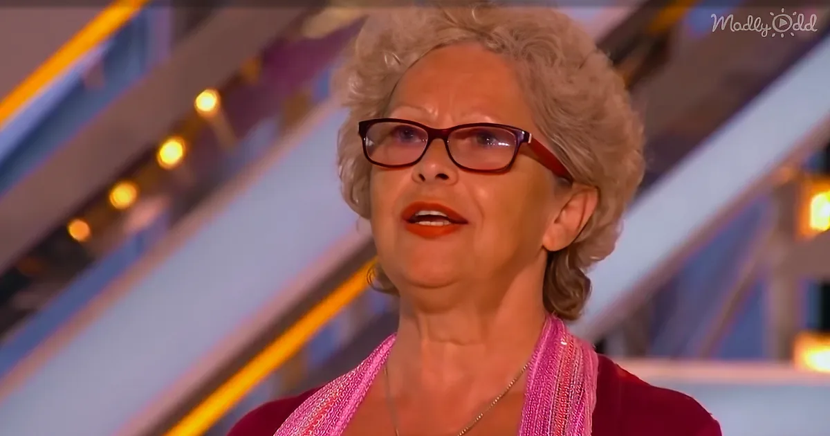 A woman with curly hair, glasses, and a pink scarf is speaking on a stage with bright lights.