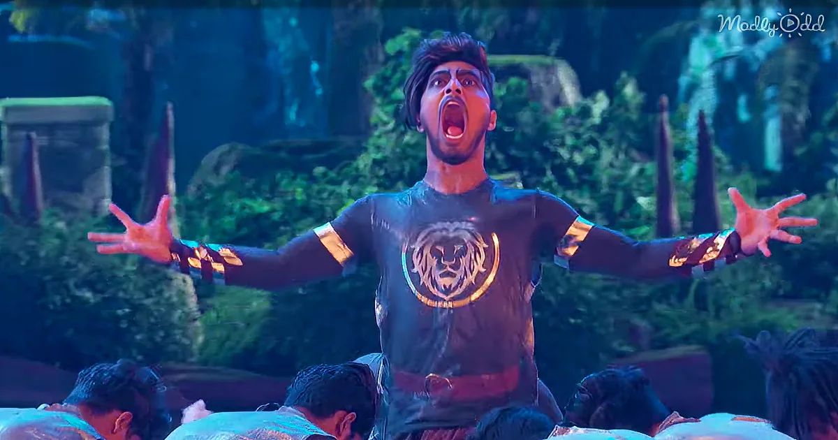 A man in a lion-themed shirt with outstretched arms and an open mouth, surrounded by a crowd and a forest-like background.