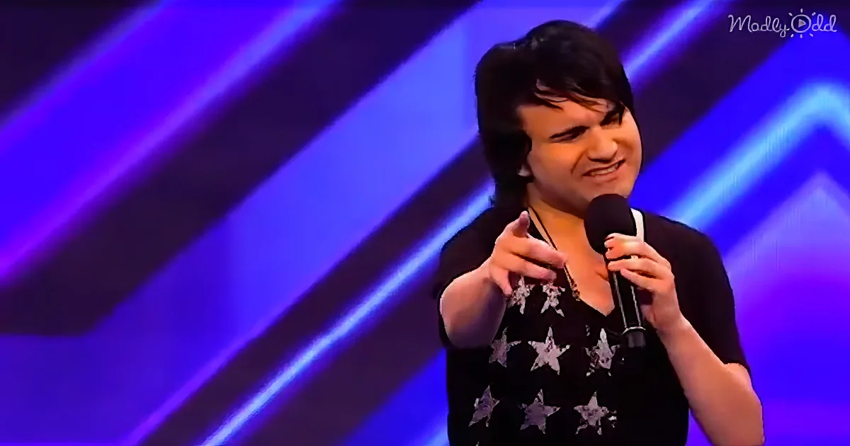 A person on stage holding a microphone, wearing a black shirt with white stars, gesturing with their left hand.