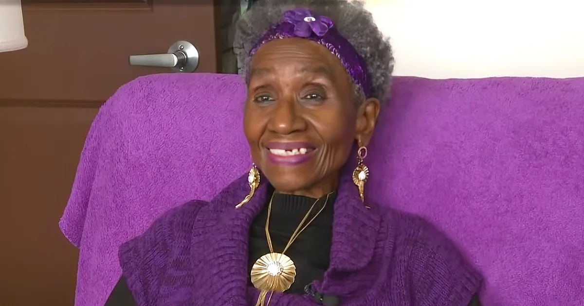 An elderly woman with purple hair accessories and jewelry smiles while sitting on a purple couch.