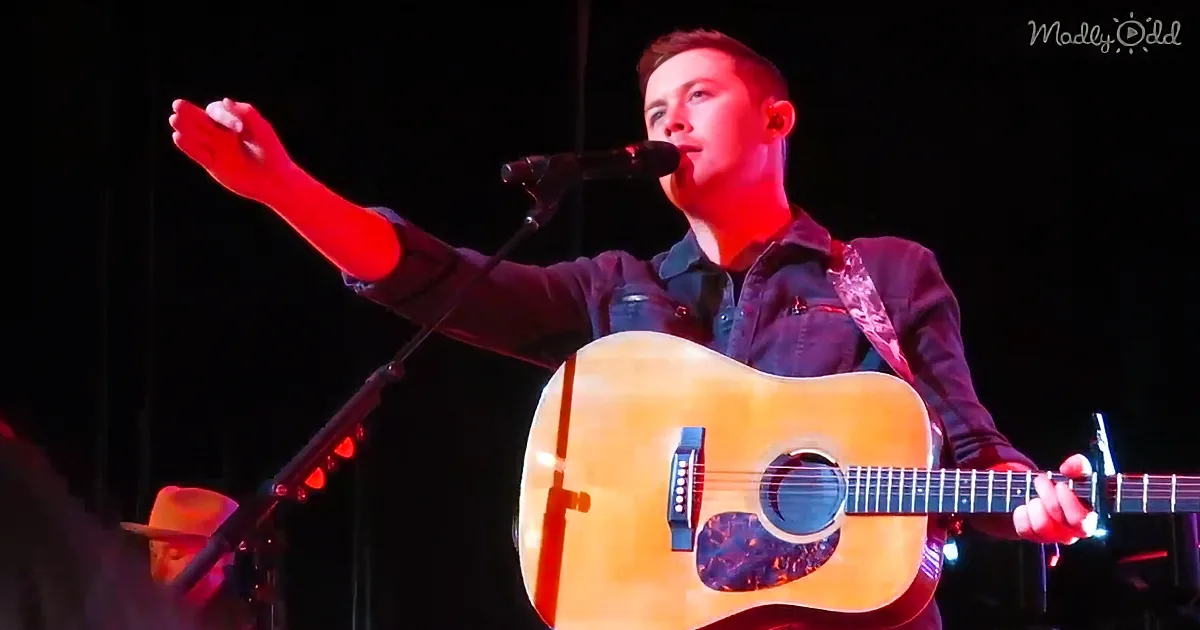 A man plays an acoustic guitar on stage, singing into a microphone with red lighting.