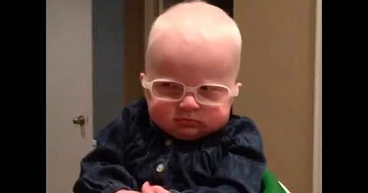 A baby wearing glasses and a dark shirt sits indoors, looking down.