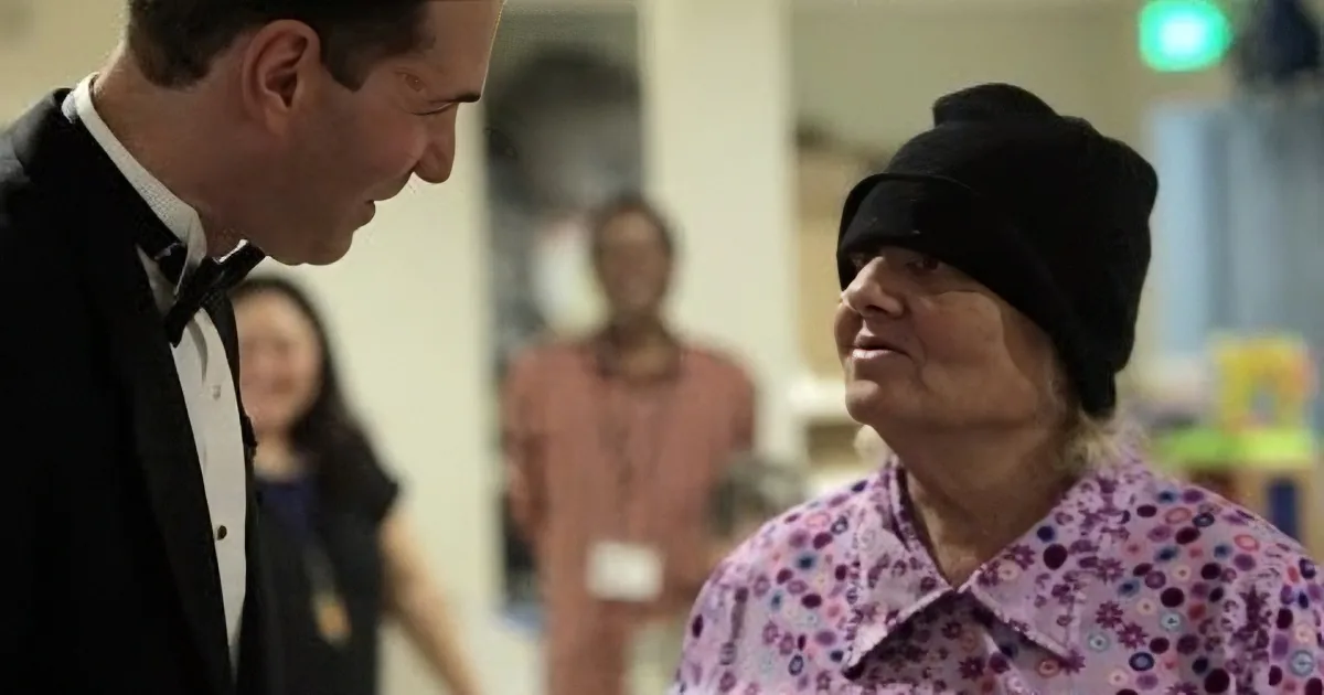 A man in a tuxedo looks at an elderly woman wearing a floral shirt and a black beanie in a room with other people.