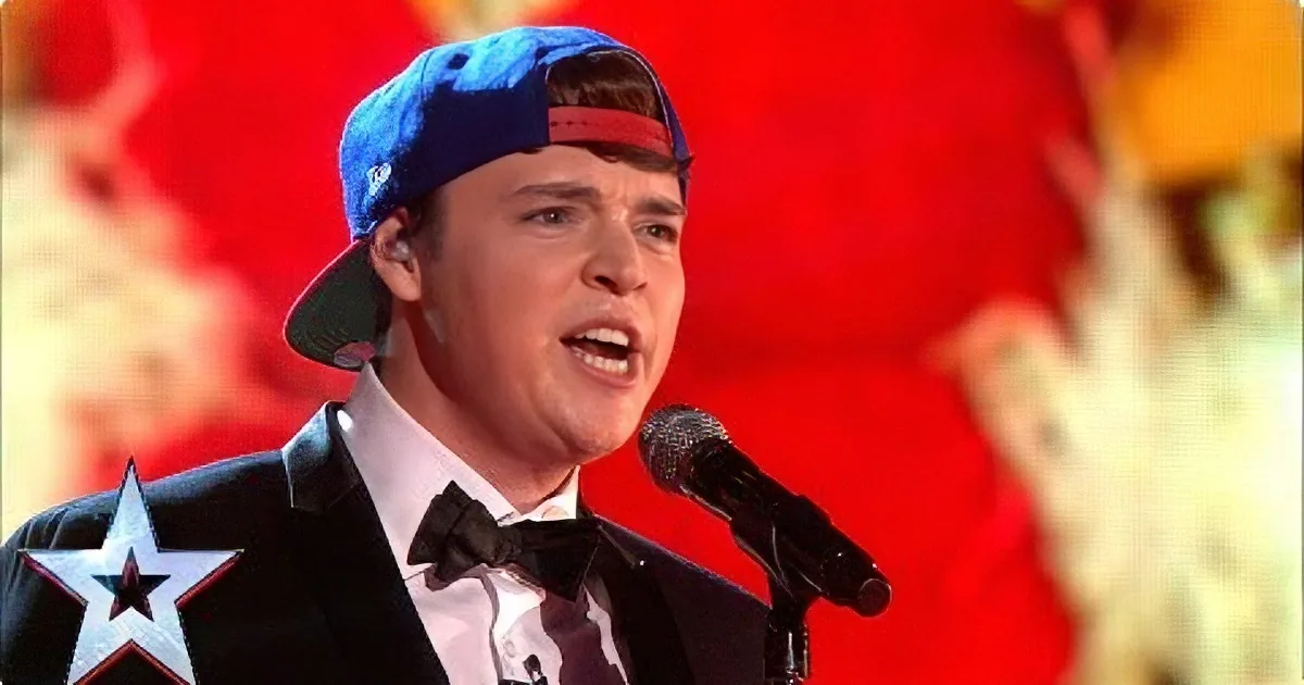 A person wearing a blue cap and a black suit with a star patch sings into a microphone on a stage with a red background.