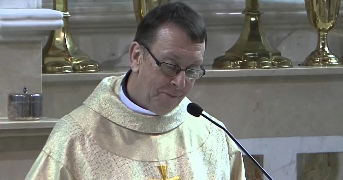A priest in a white robe speaks into a microphone, wearing glasses, in a church setting.
