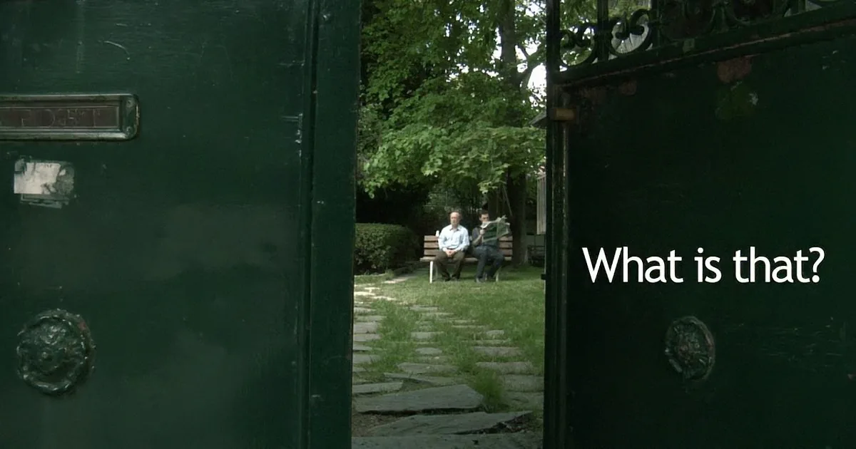 A man sits on a bench in a garden, framed by a green gate with a letterbox and ornate handles.
