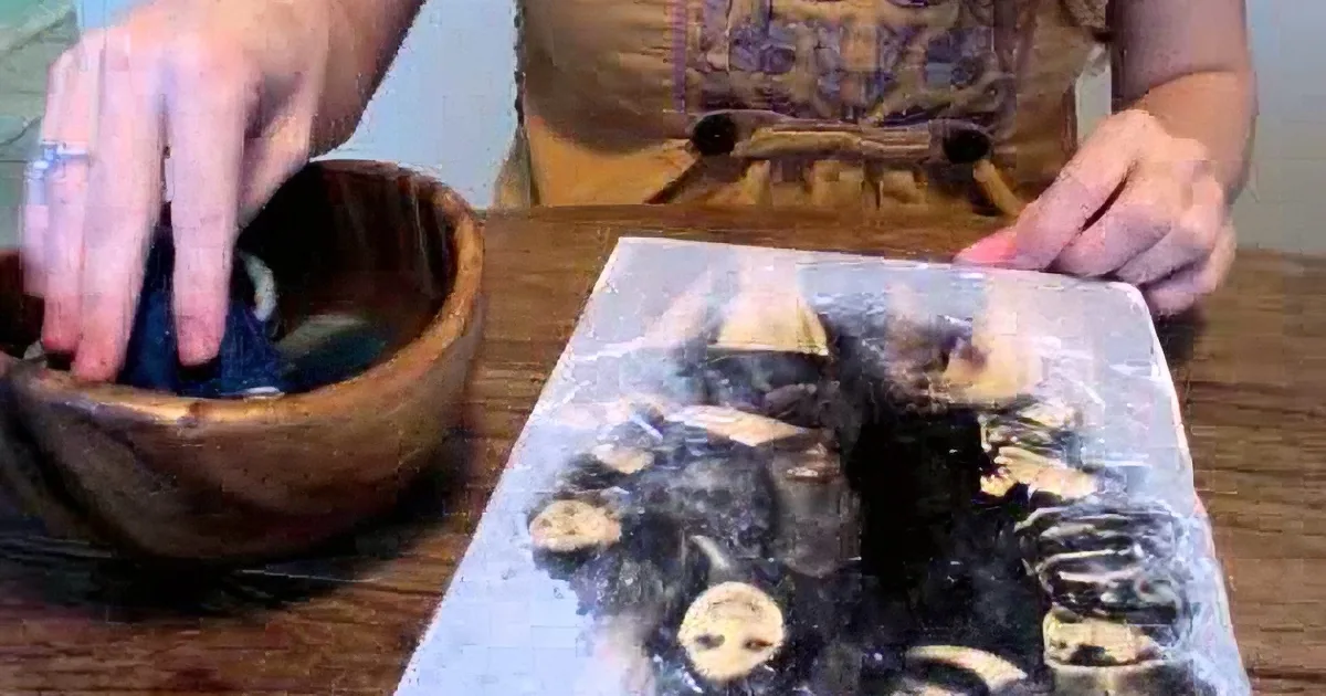 A person prepares a dish with a wooden bowl and a plastic bag containing black and white ingredients on a wooden table.