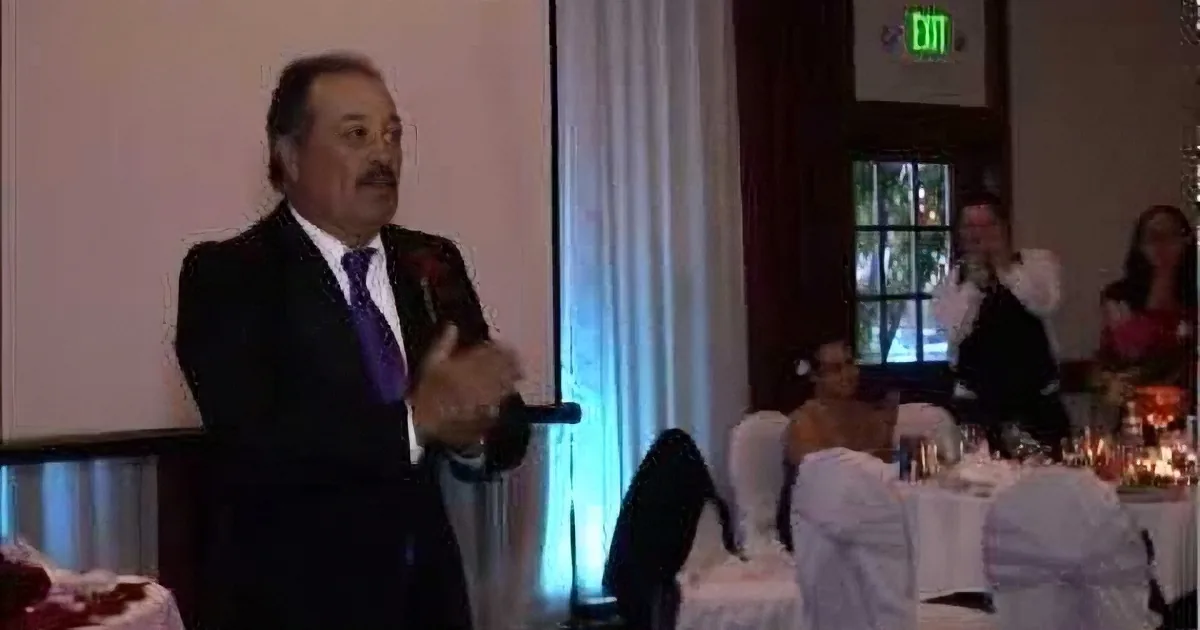 A man in a suit and tie is giving a speech at a formal event with tables and guests in the background.