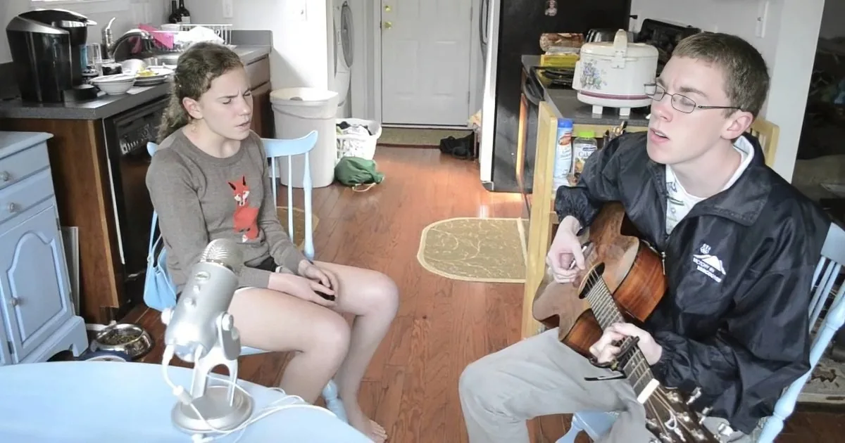 A man plays guitar while a woman sings into a microphone in a kitchen setting.
