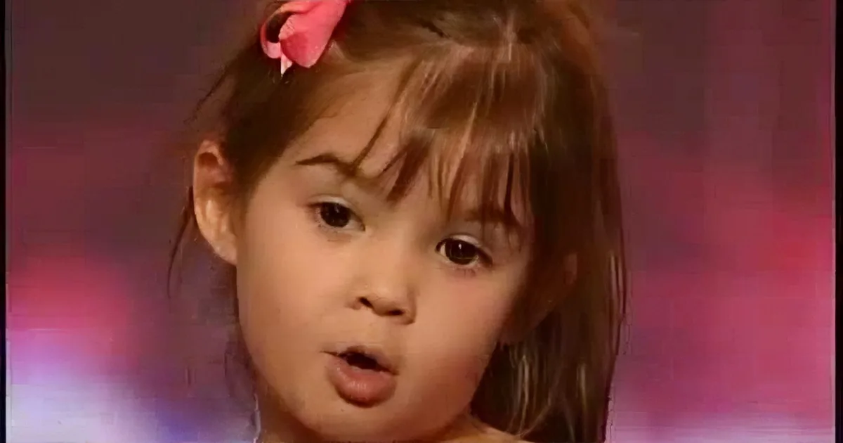 A young girl with brown hair and a pink bow, wearing a red outfit, appears to be speaking or singing.