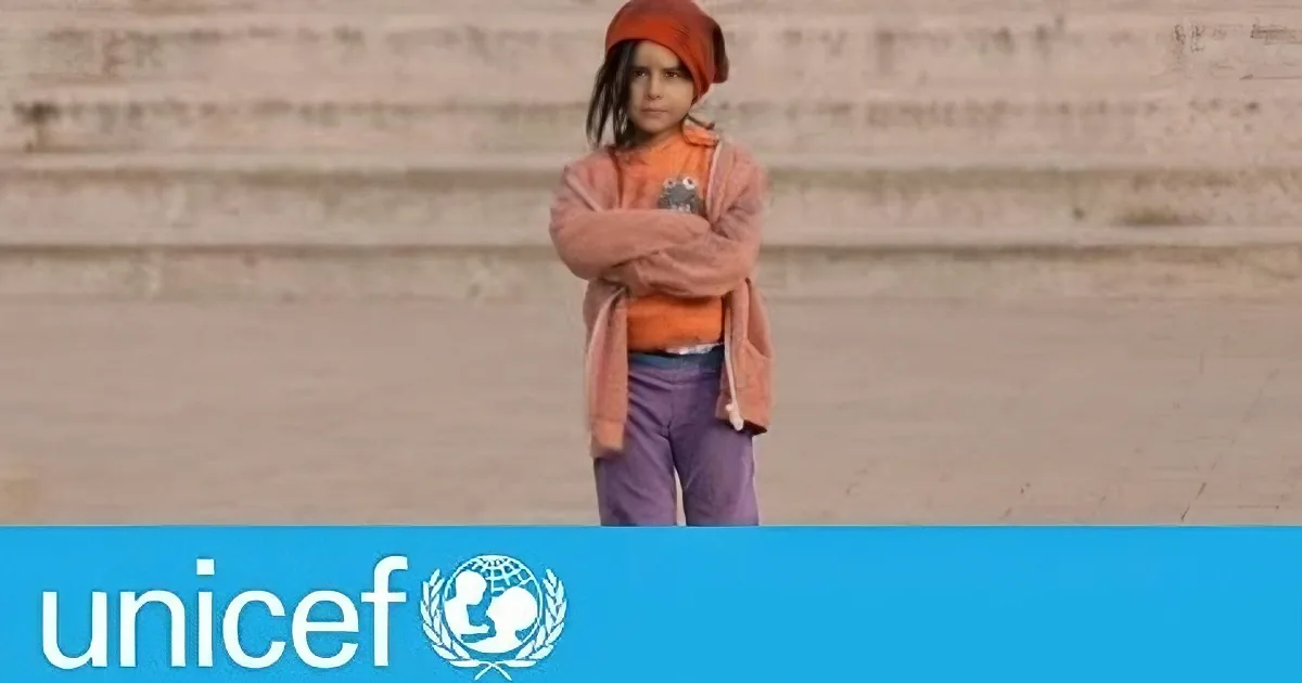 A child stands in a barren landscape, wearing an orange hat and jacket, with arms crossed, in front of a UNICEF logo.