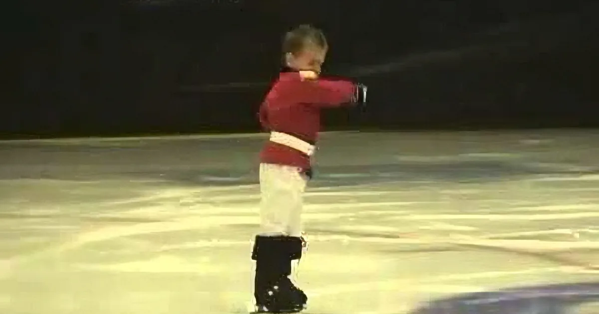 A child in a red sweater and white pants is skating on ice, holding a stick.
