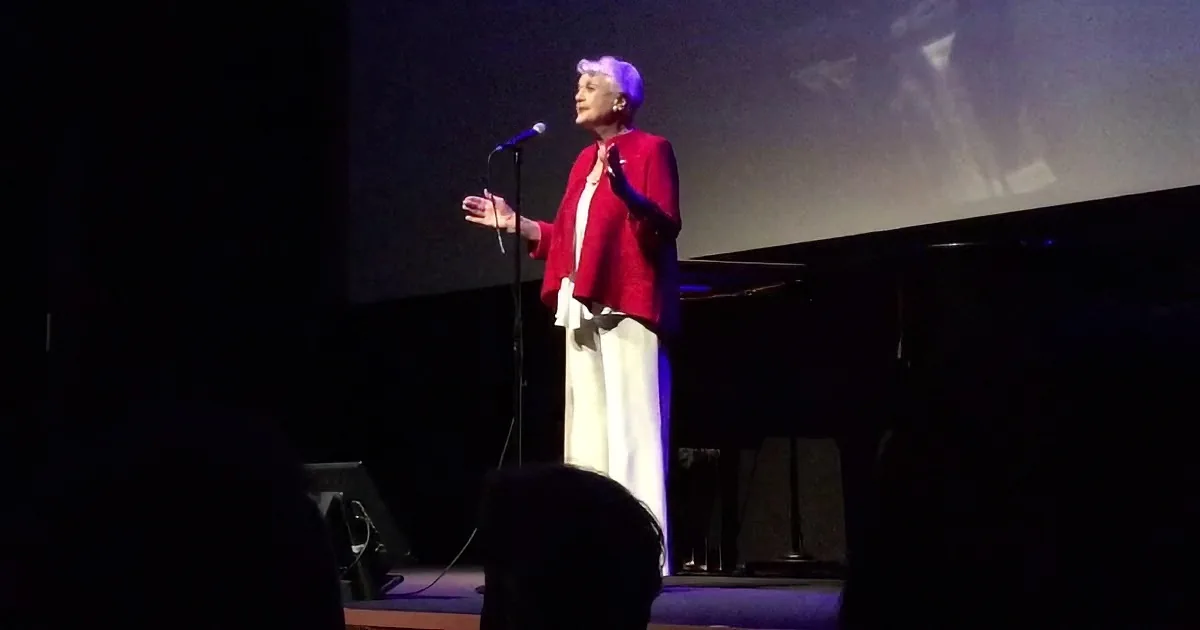 A woman in a red jacket and white pants stands on stage, gesturing with her hands near a microphone.