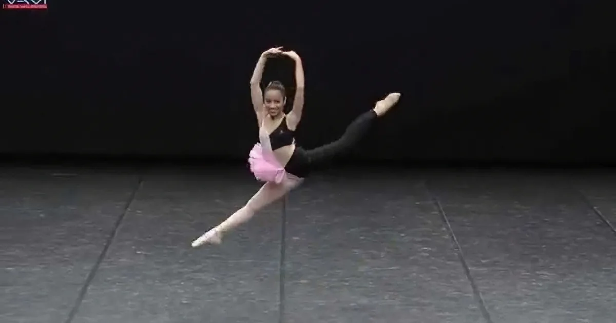 A ballerina in a pink tutu and black leotard performs a graceful split on a dark stage.