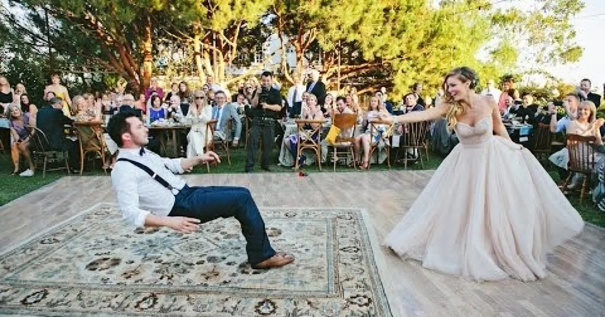 A bride in a blush gown dances with a groom in a white shirt and suspenders on a rug in front of a seated audience.