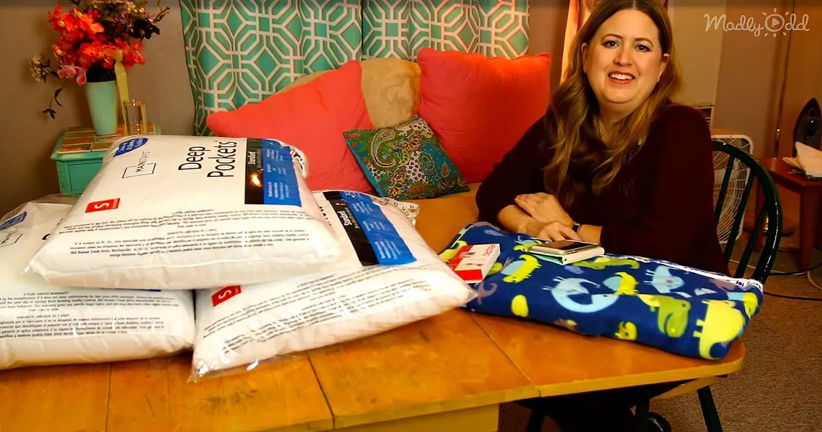 A woman sits at a table with pillows, a blanket, and a box, smiling in a cozy room.
