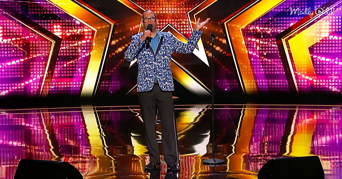 A man in a patterned blazer and glasses stands on stage with a microphone, gesturing with his hands.