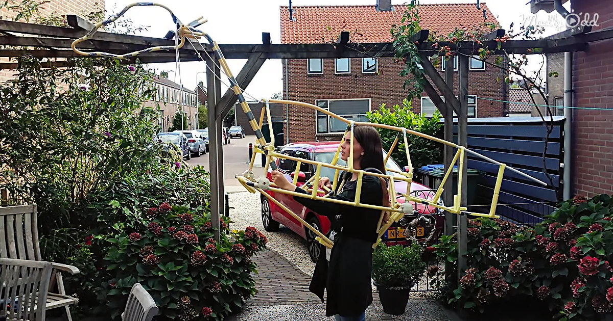 A woman holds a yellow and black structure resembling a bridge or sculpture in a garden setting.
