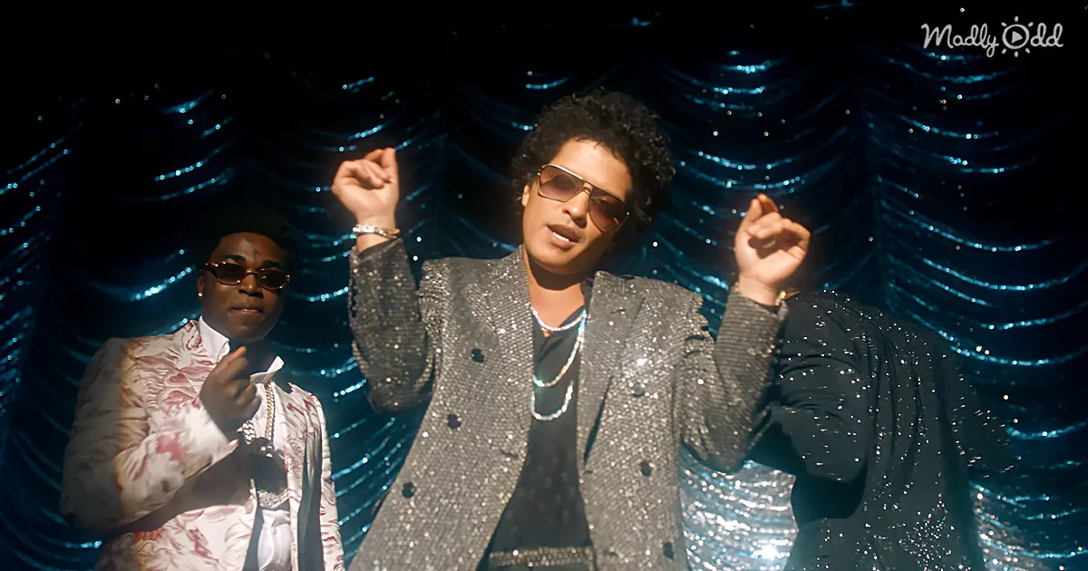 A man in a glittery suit raises his fists, wearing sunglasses, with another person in a patterned jacket behind him.