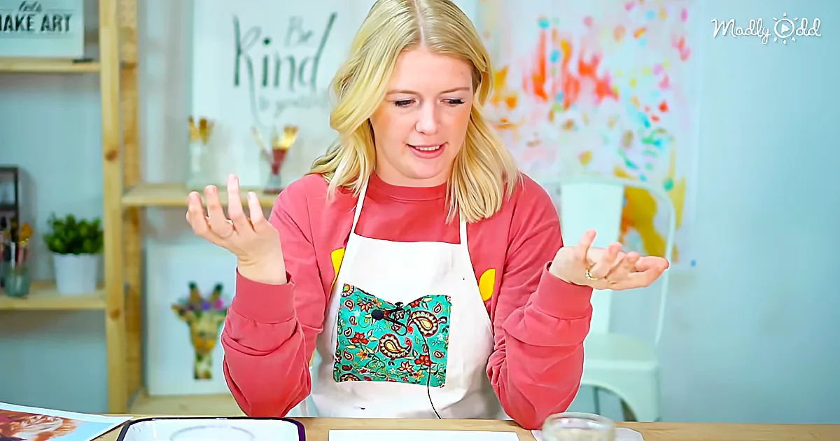 A woman in a red sweater and apron gestures with her hands, standing in front of a colorful art setup.