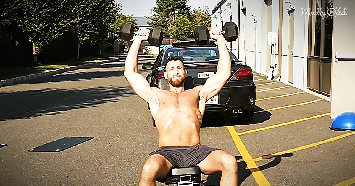 A shirtless man is doing a seated dumbbell press in a parking lot with a car and building in the background.